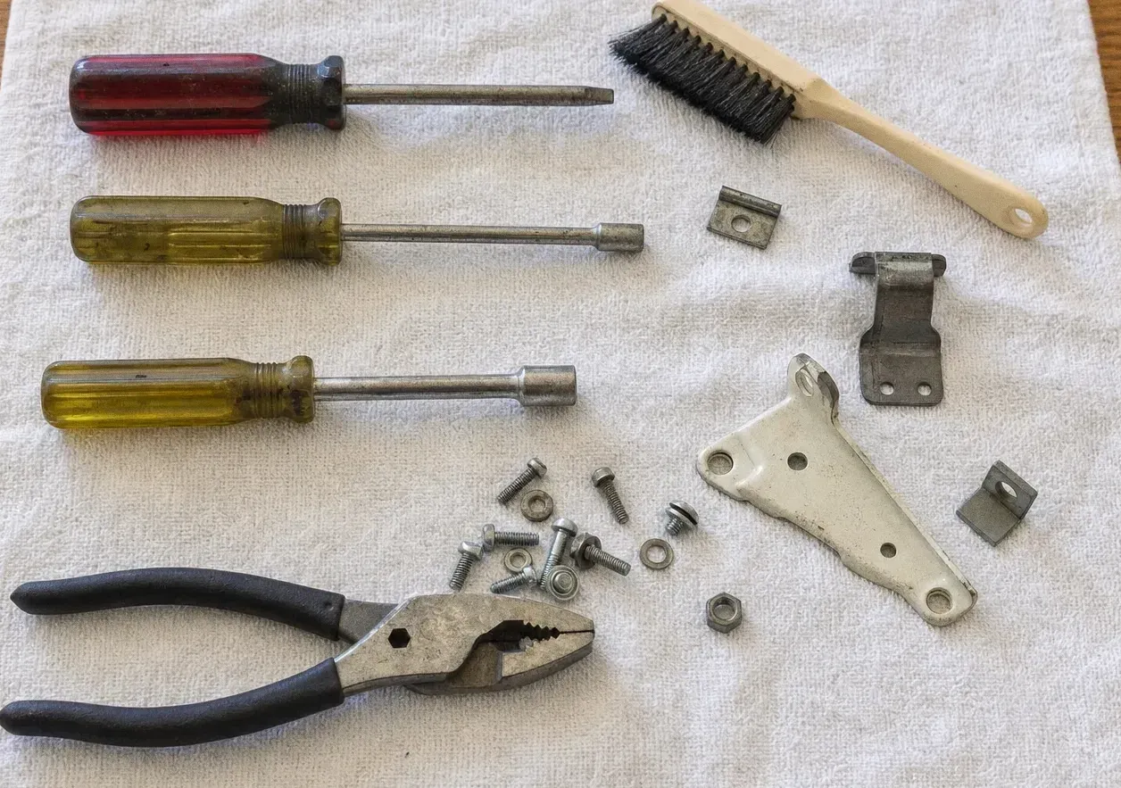 Screwdrivers, nut drivers, a wire brush and slip-joint pliers laid out on a towel with hinge brackets, bolts and washers