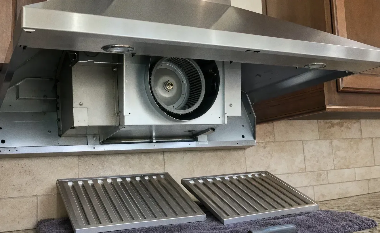 A range hood canopy opened to show the blower wheel inside, with two baffle filters standing on the counter against a tiled backsplash